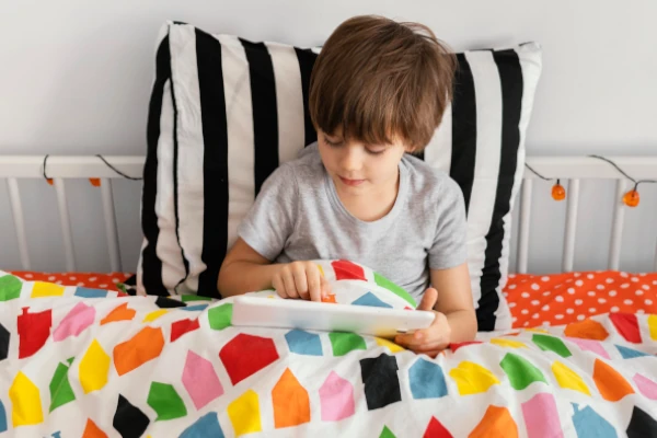 Chłopiec z tabletem w łóżku pod kolorową kołdrą 